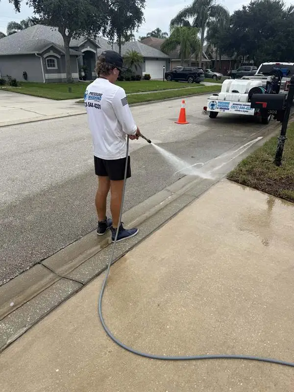 Technician pressure washing a curbside sidewalk with the wash trailer nearby in Rockledge, FL